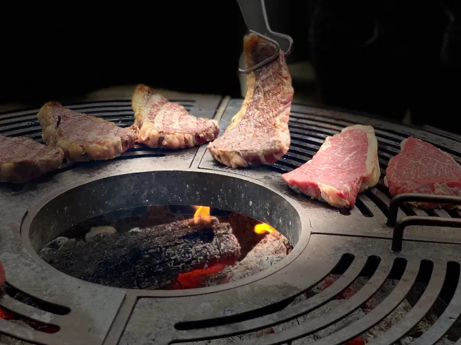 Pièces de bœuf sur la plancha, braises visibles au centre, et morceaux crus persillés en attente.